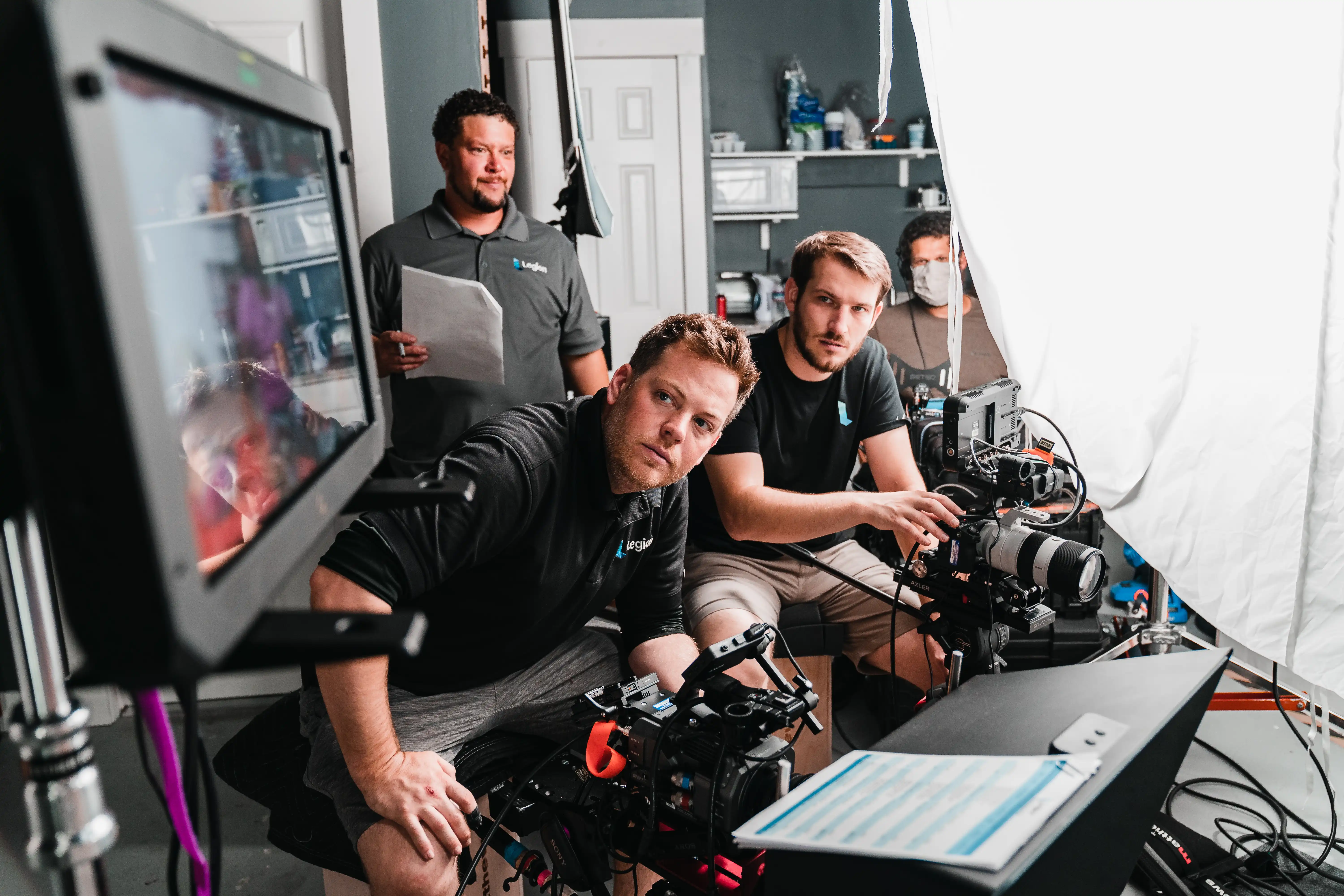 A group of camera operators, a director, and a sound operator all looking at a monitor on set. The team is focused on the monitor as they review the footage or setup.